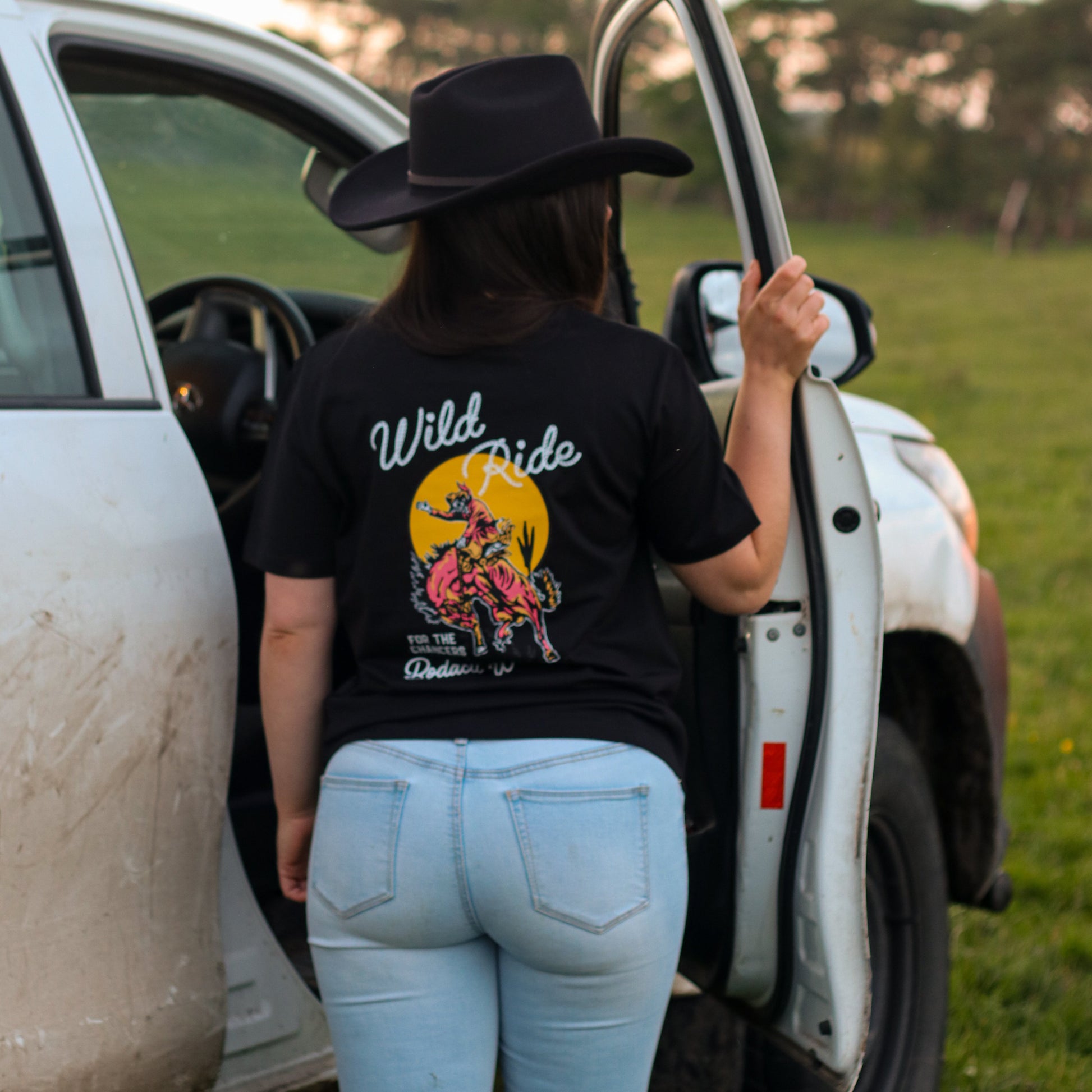A woman wearing a black cowboy hat and a black western graphic t-shirt with 'Wild Ride' printed on the back, standing next to an open white truck door in an outdoor grassy field.