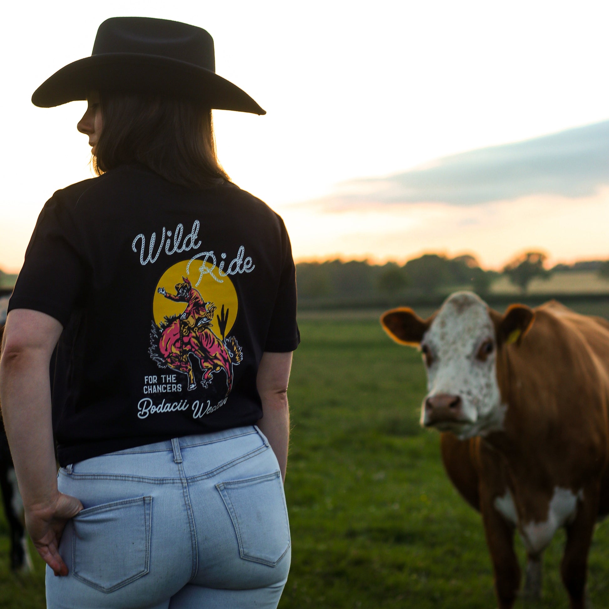 A cowgirl wearing a black cowboy hat and a black graphic t-shirt with a 'Wild Ride' design, standing in a grassy field with a brown and white cow next to her at sunset.