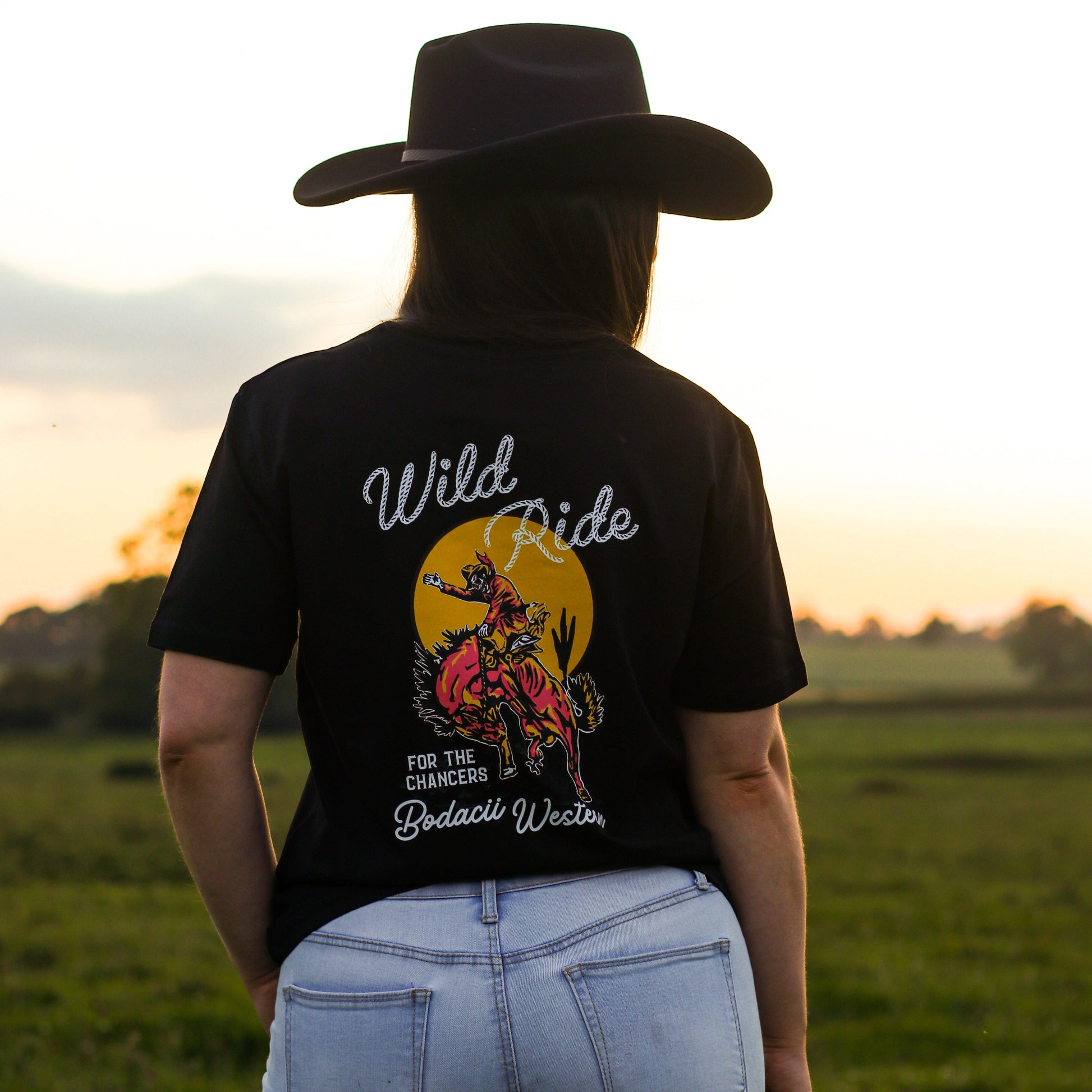 A cowgirl viewed from behind wearing a black graphic t-shirt with a 'Wild Ride' bodacii western design, paired with light blue jeans and a black cowboy hat, standing in a grassy field at sunset.