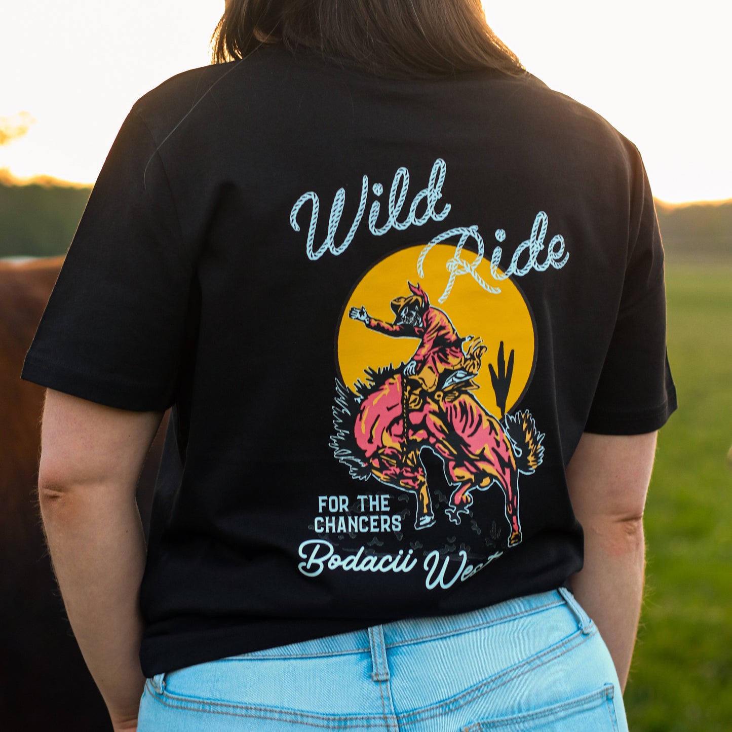 A woman wearing a black graphic t-shirt and a black cowboy hat, viewed from behind in an outdoor field at sunset. The back of the t-shirt features a colourful graphic of a cowboy riding a bucking bronc in front of a yellow sun, with the text 'Wild Ride' and 'FOR THE CHANCERS Bodacii western